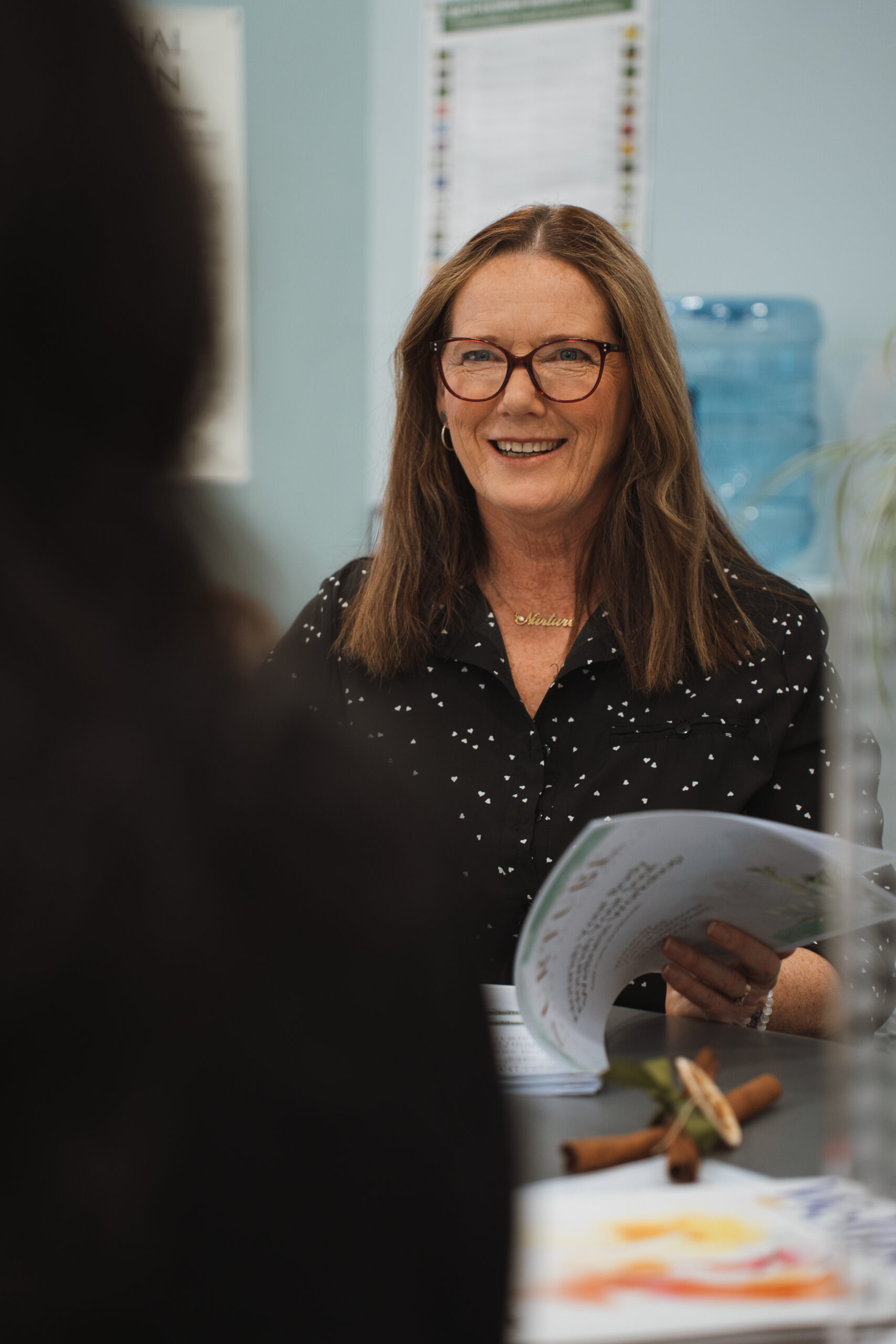 Louise from Nurture with Louise, a smiling naturopath, holds a wellness booklet while seated at a table, ready to consult.